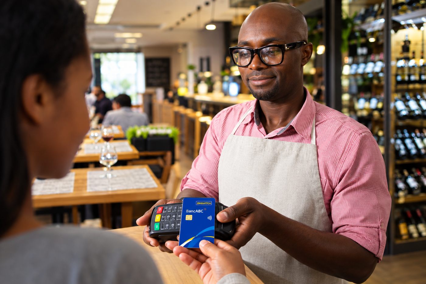 A waiter holds a card terminal while a customer pays by card at a restaurant table